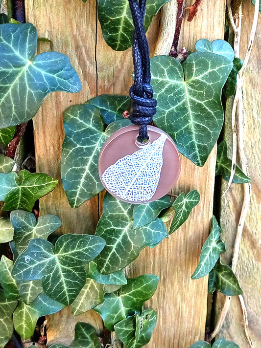 Warm brown round pendant with cream leaf skeleton design against ivy covered wood in natural daylight
