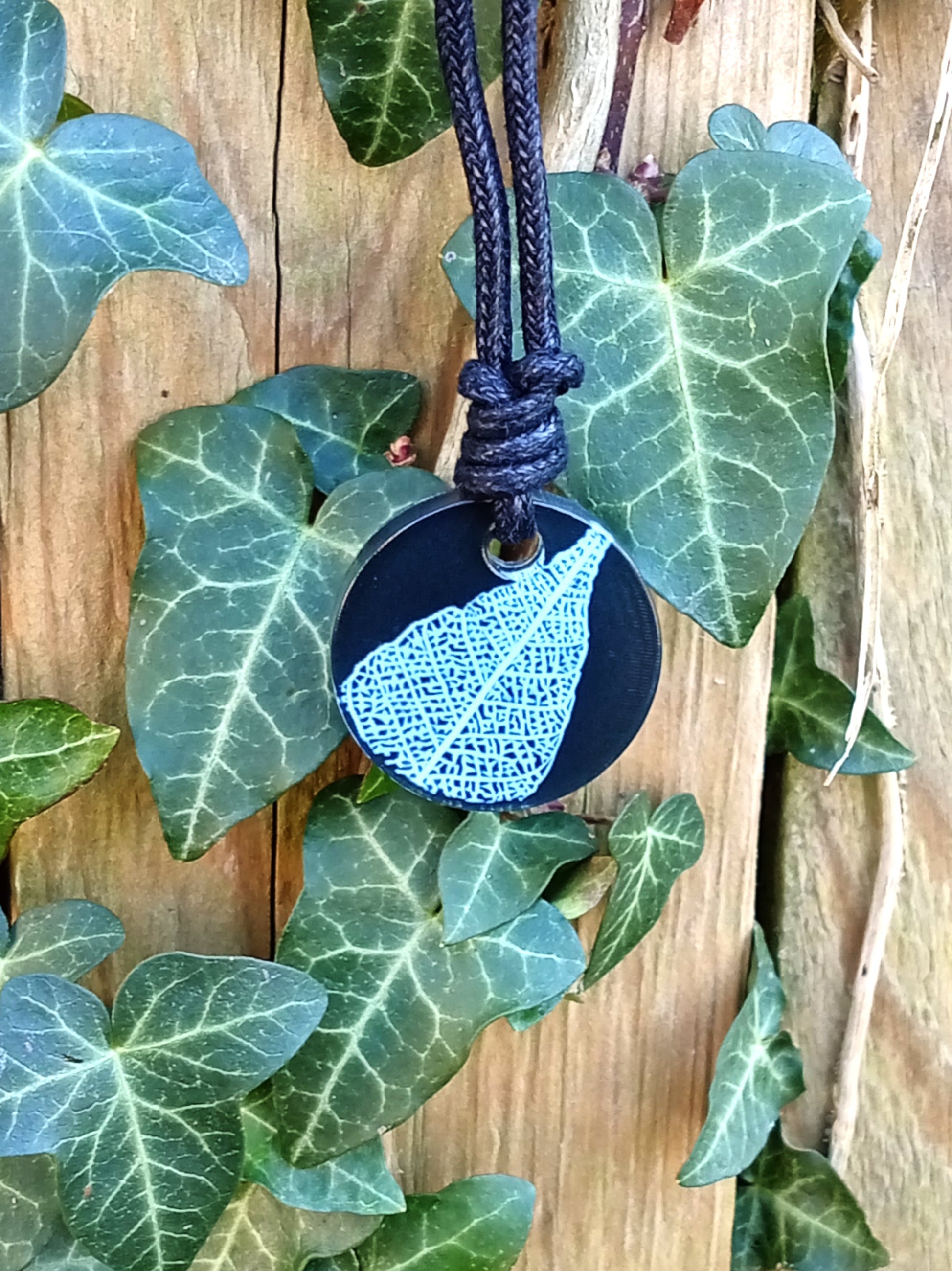 Black round pendant with white leaf skeleton design against ivy covered wood in natural daylight
