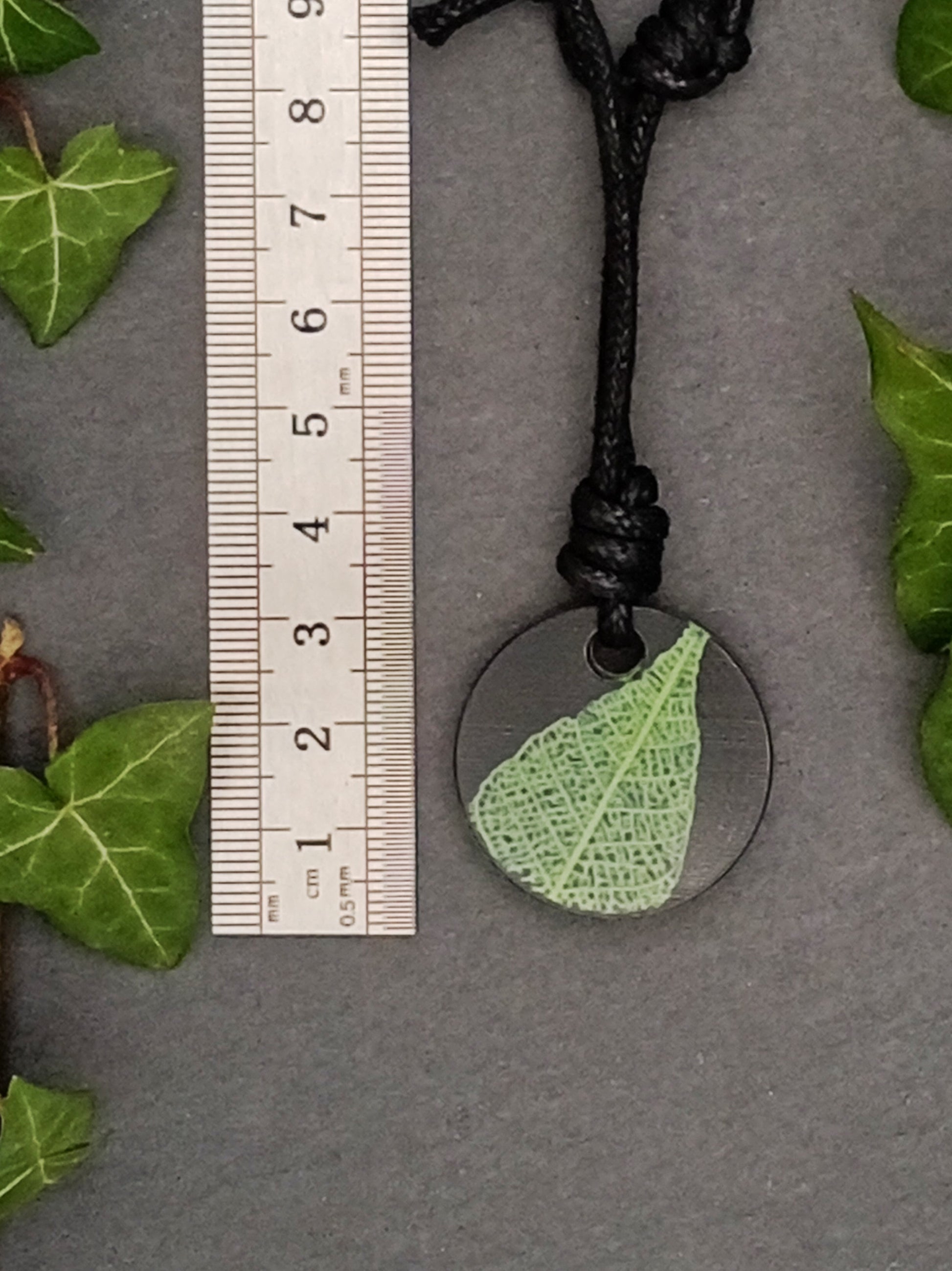 Black round pendant with bright green leaf skeleton design shown next to a ruler displaying centimetres for scale