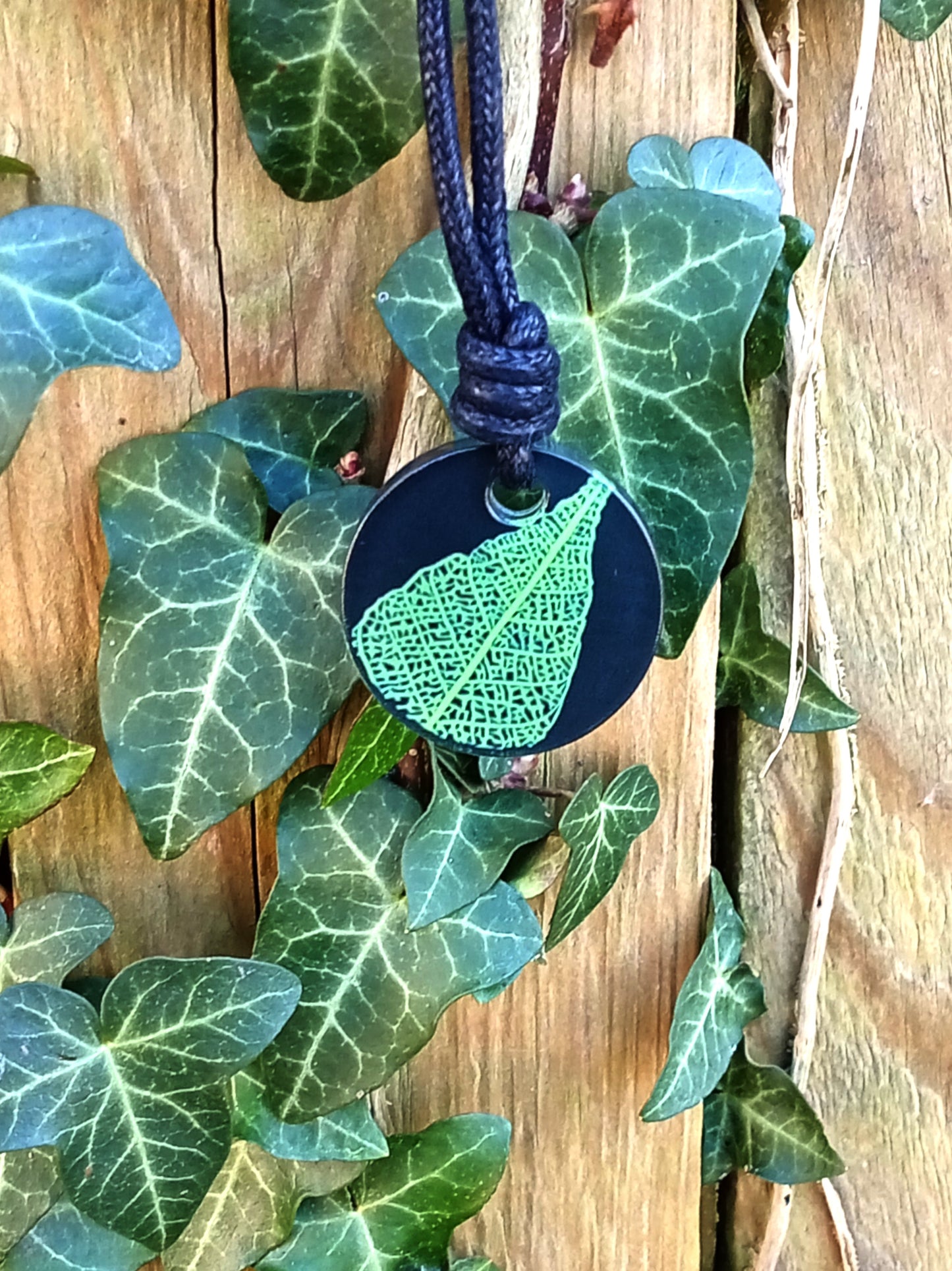 Black round pendant with bright green leaf skeleton design against ivy covered wood in natural daylight