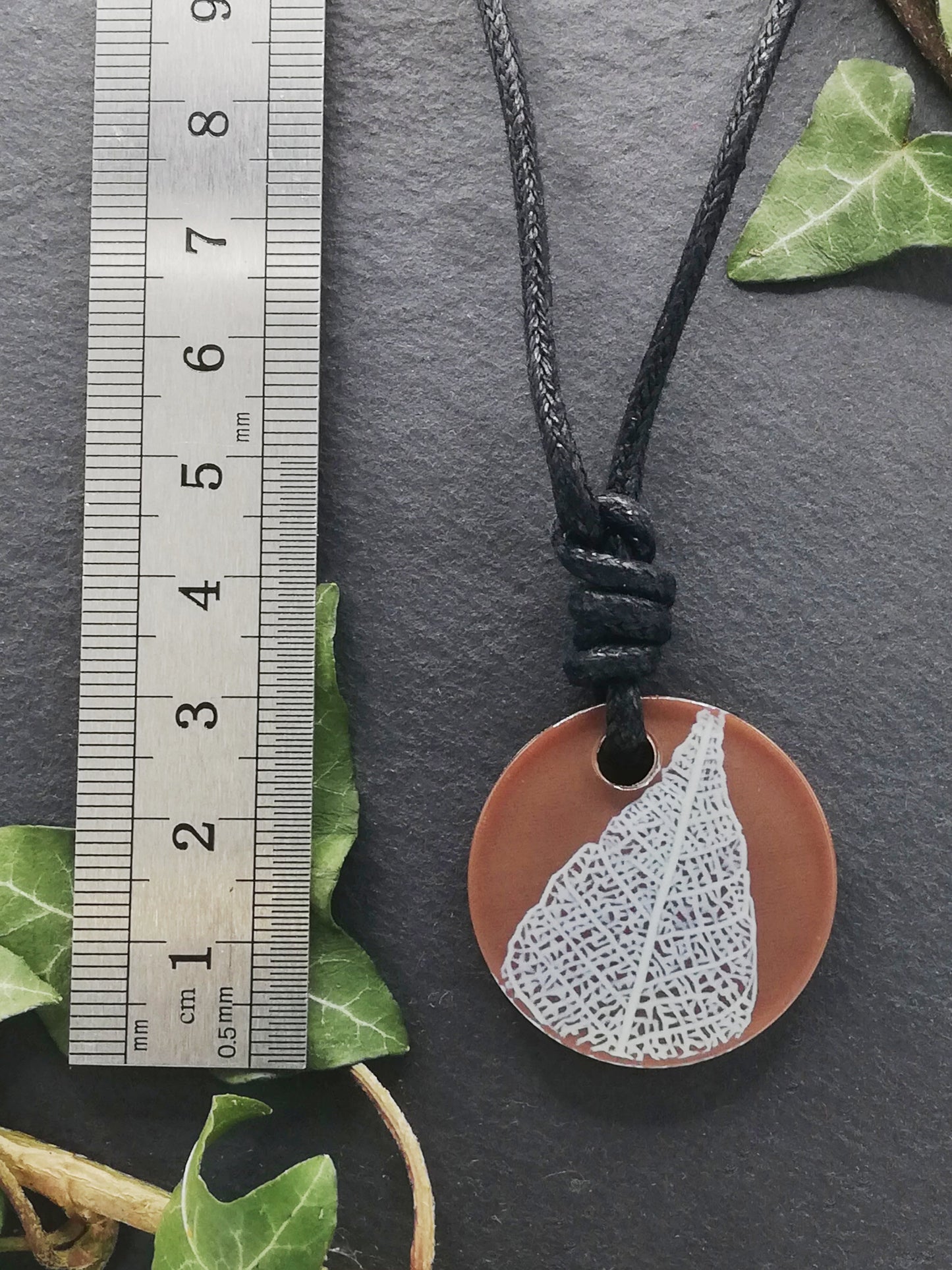 Warm brown round pendant with cream leaf skeleton design shown next to a ruler displaying centimetres for scale