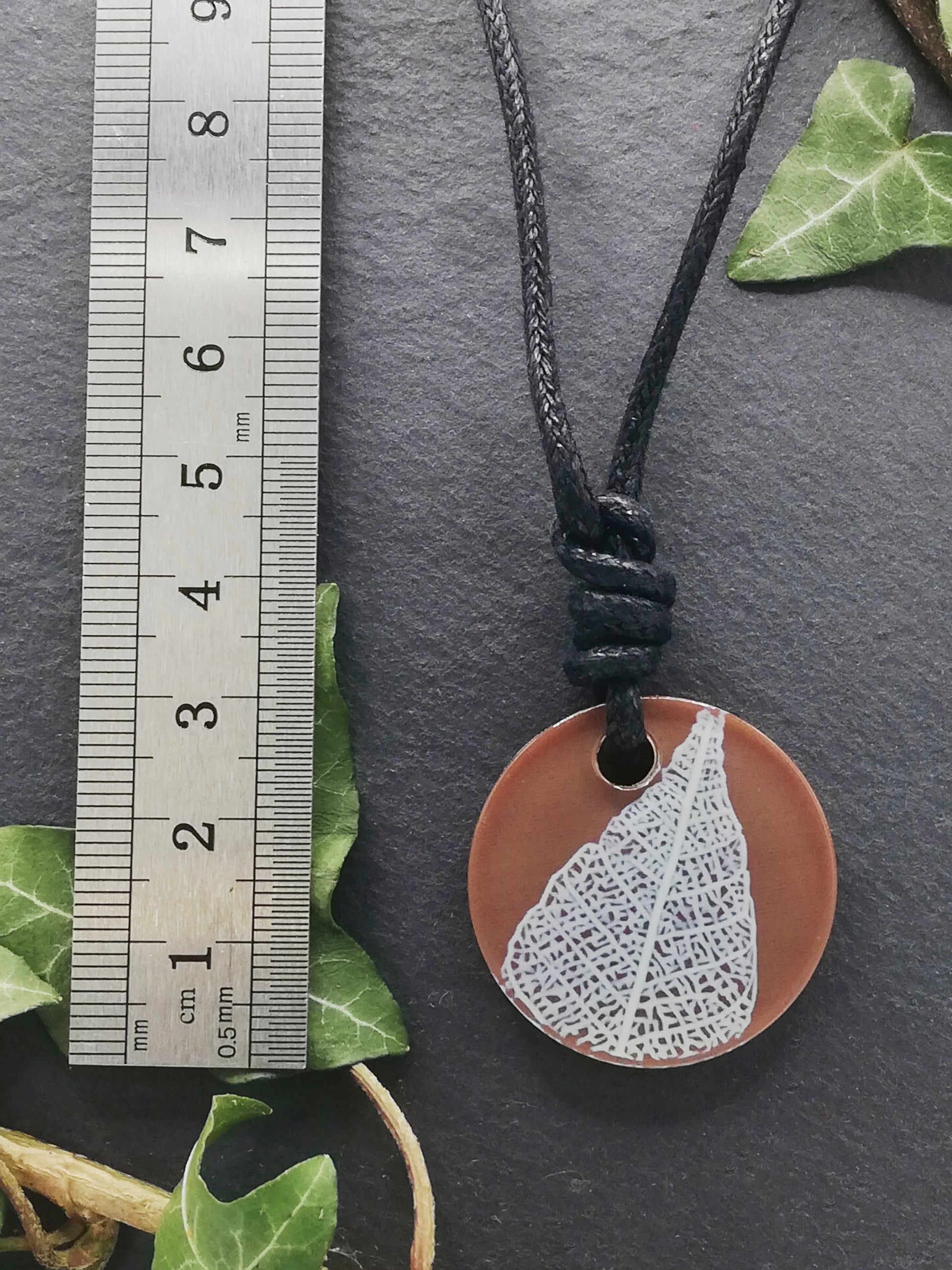 Warm brown round pendant with cream leaf skeleton design shown next to a ruler displaying centimetres for scale
