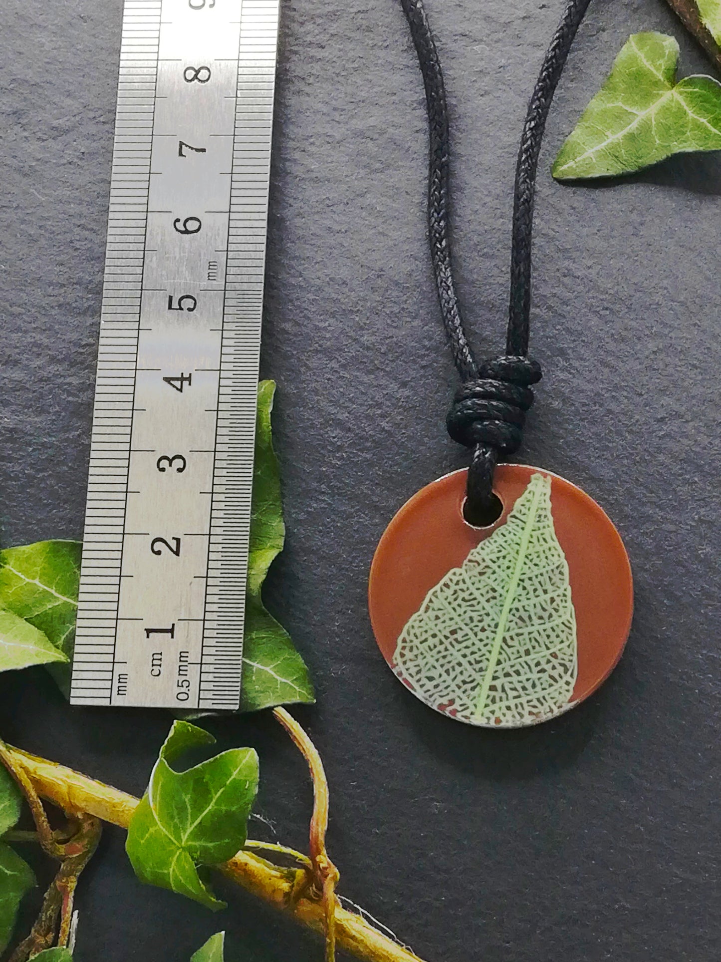 Warm brown round pendant with pale green leaf skeleton design shown next to a ruler displaying centimetres for scale