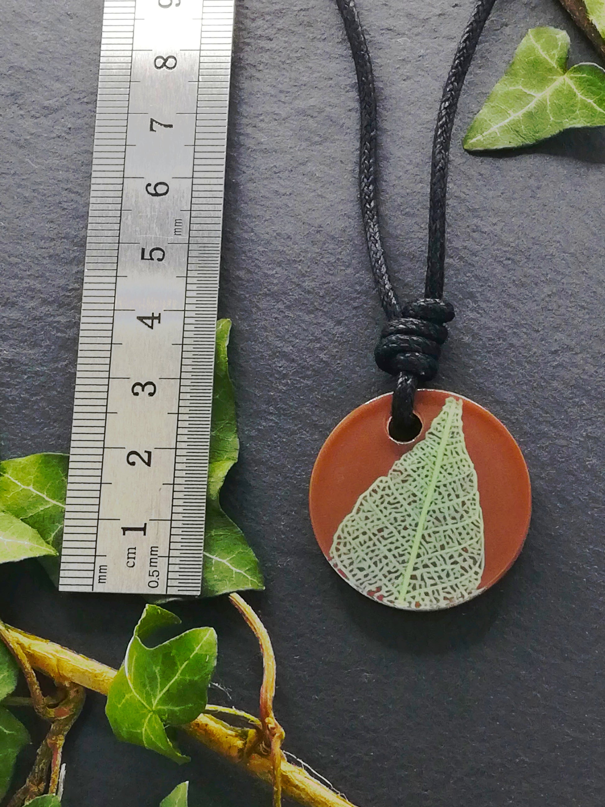 Warm brown round pendant with pale green leaf skeleton design shown next to a ruler displaying centimetres for scale
