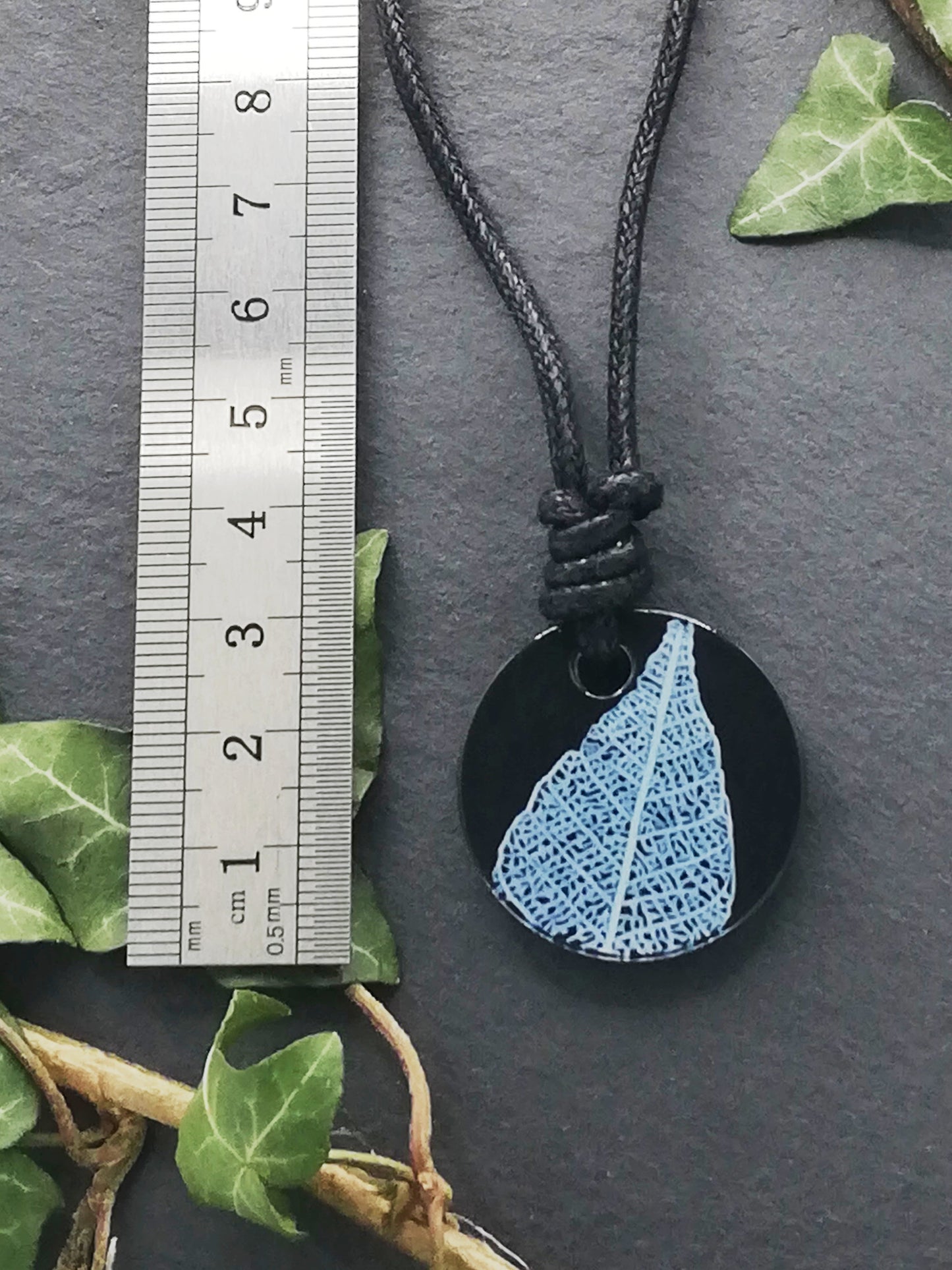 Black round pendant with white leaf skeleton design shown next to a ruler displaying centimetres for scale