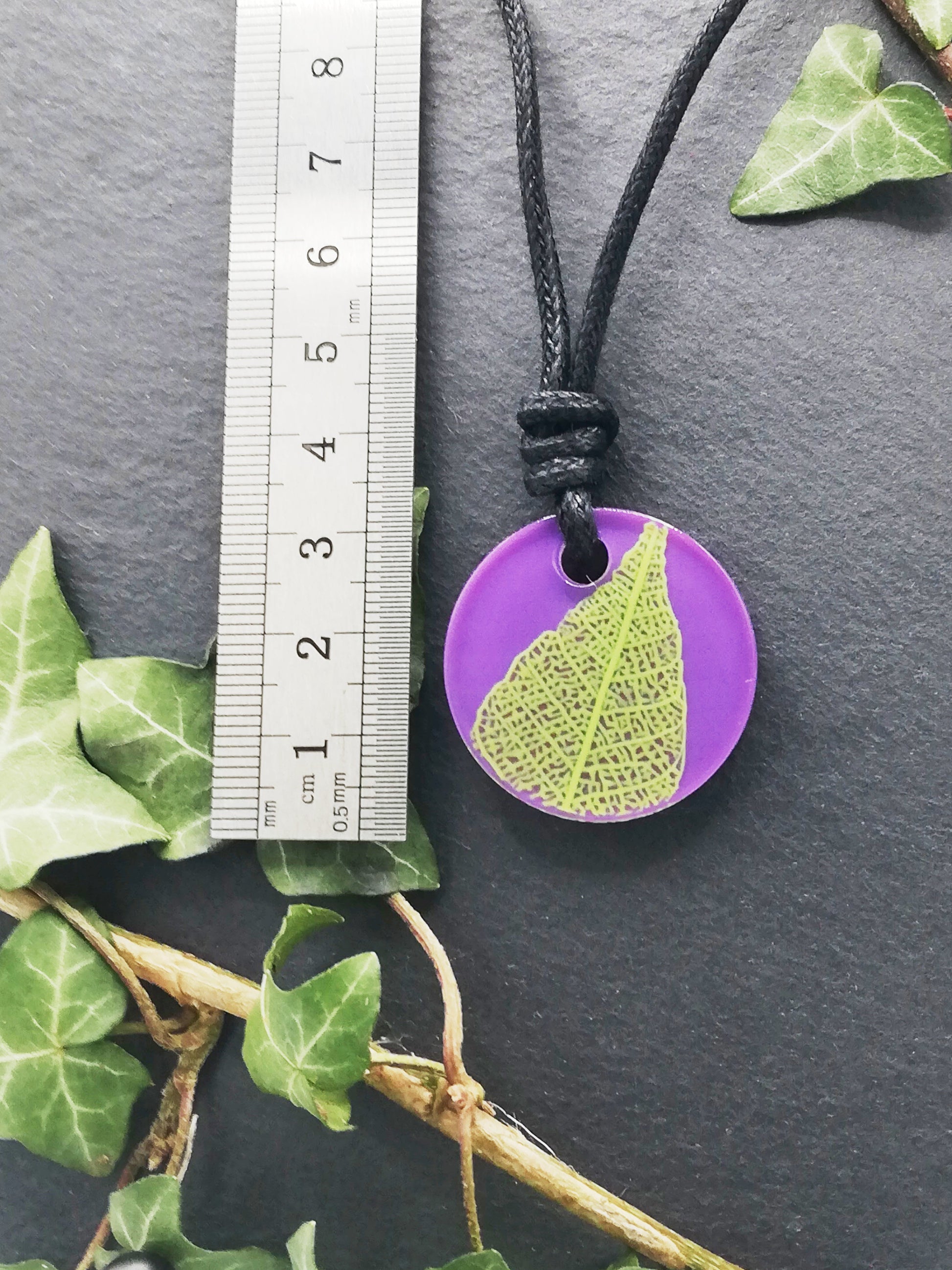 Purple round pendant with bright green leaf skeleton design shown next to a ruler displaying centimetres for scale