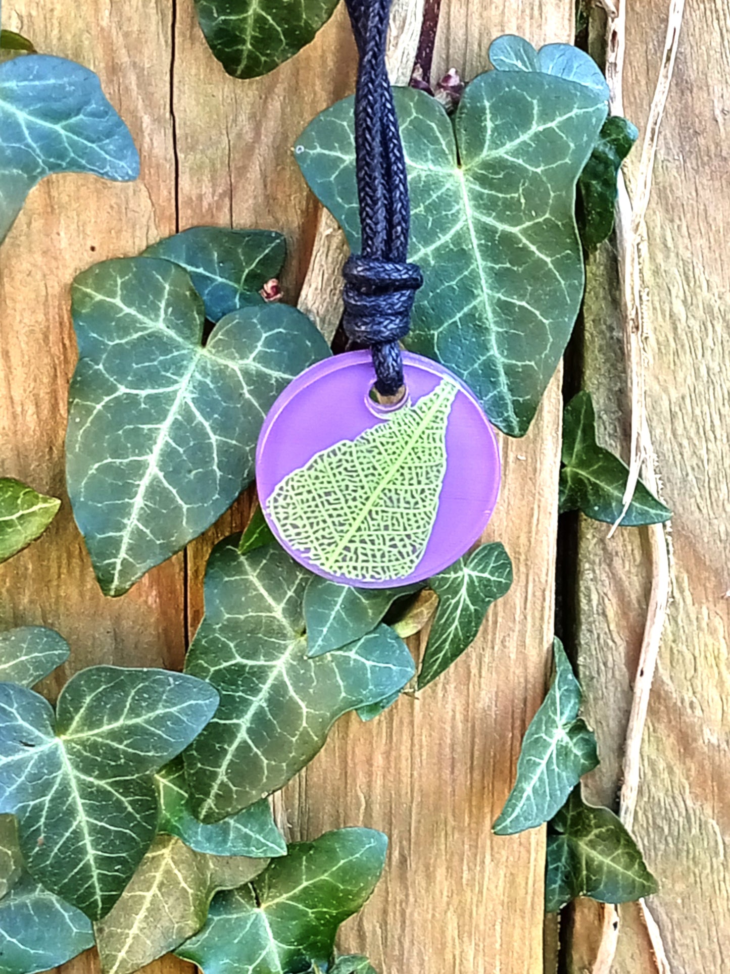 Purple round pendant with bright green leaf skeleton design against ivy covered wood in natural daylight