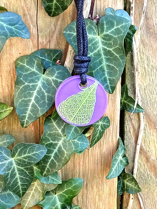 Purple round pendant with bright green leaf skeleton design against ivy covered wood in natural daylight