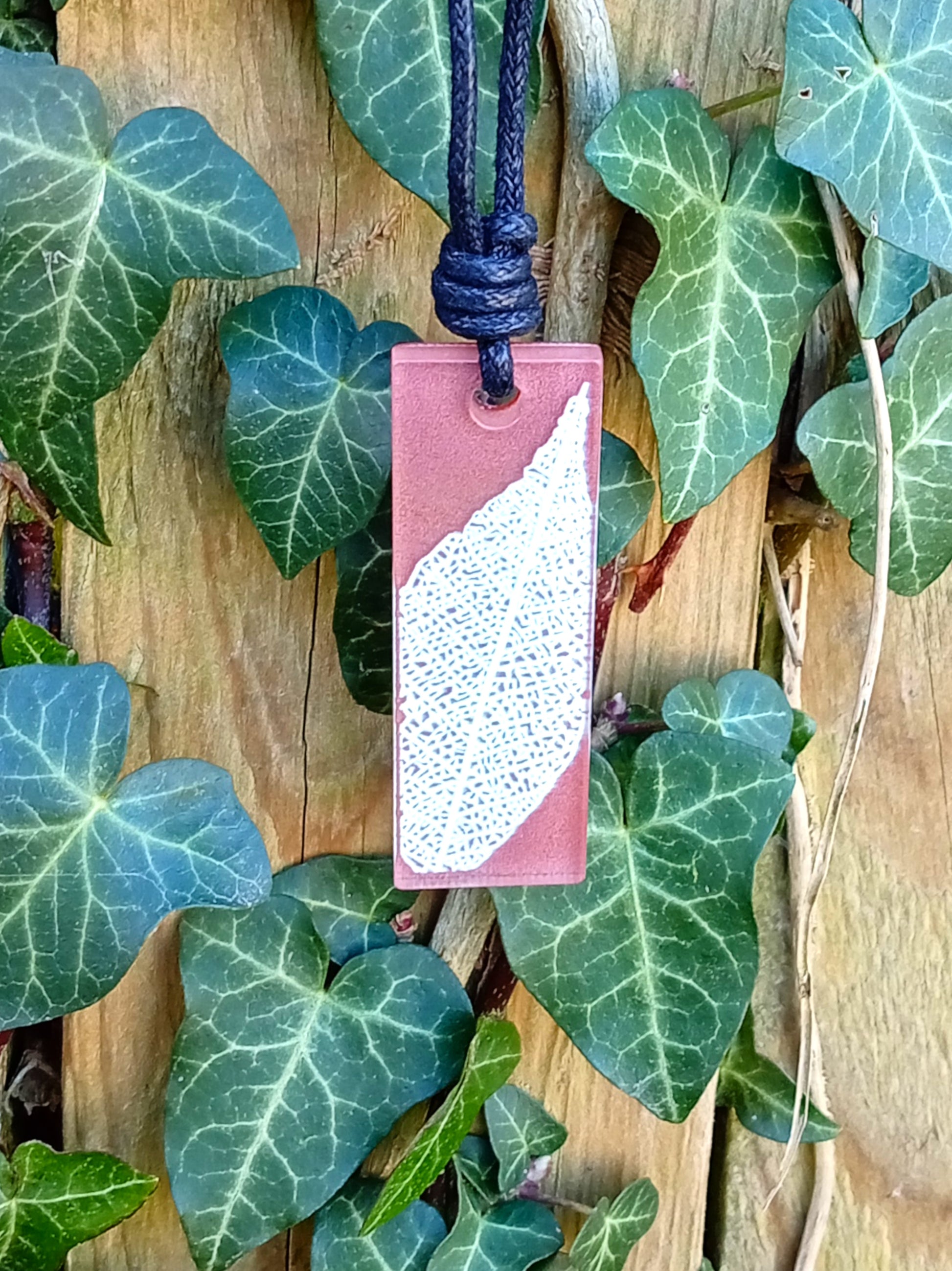Warm brown rectangular pendant with cream leaf skeleton design against ivy covered wood in natural daylight