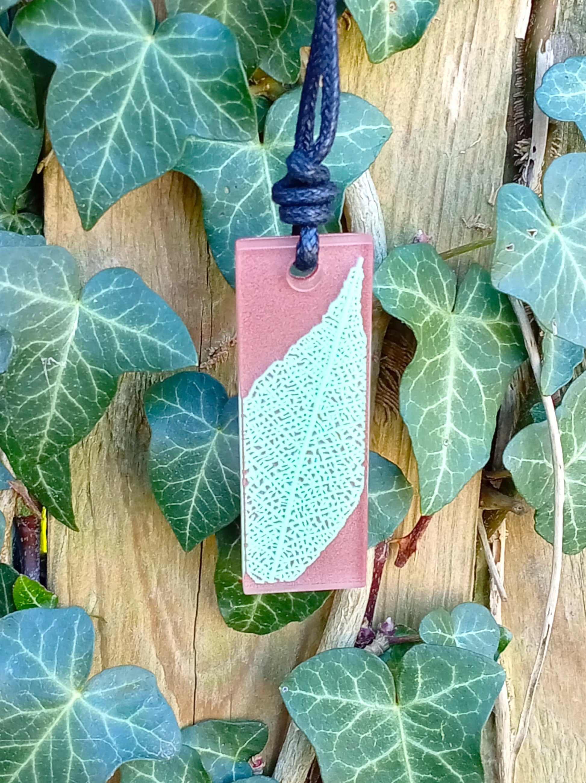 Warm brown rectangular pendant with pale green leaf skeleton design against ivy covered wood in natural daylight