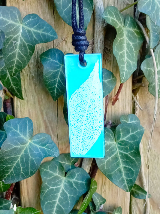 Light blue rectangular pendant with white leaf skeleton design against ivy covered wood in natural daylight