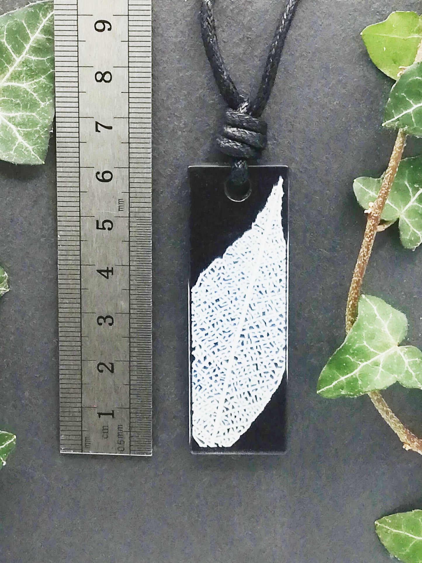 Black rectangular pendant with white leaf skeleton design shown next to a ruler displaying centimetres for scale
