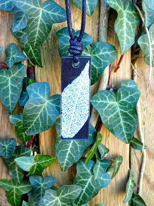 Black rectangular pendant with white leaf skeleton design against ivy covered wood in natural daylight