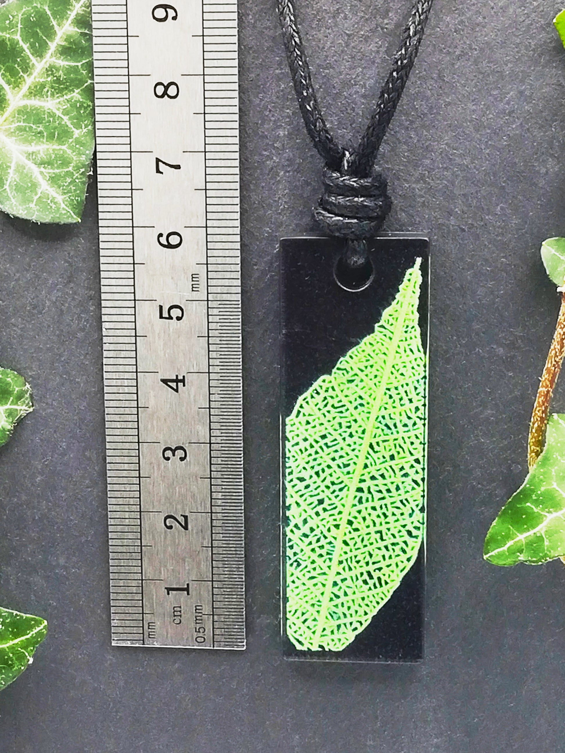 Black rectangular pendant with bright green leaf skeleton design shown next to a ruler displaying centimetres for scale