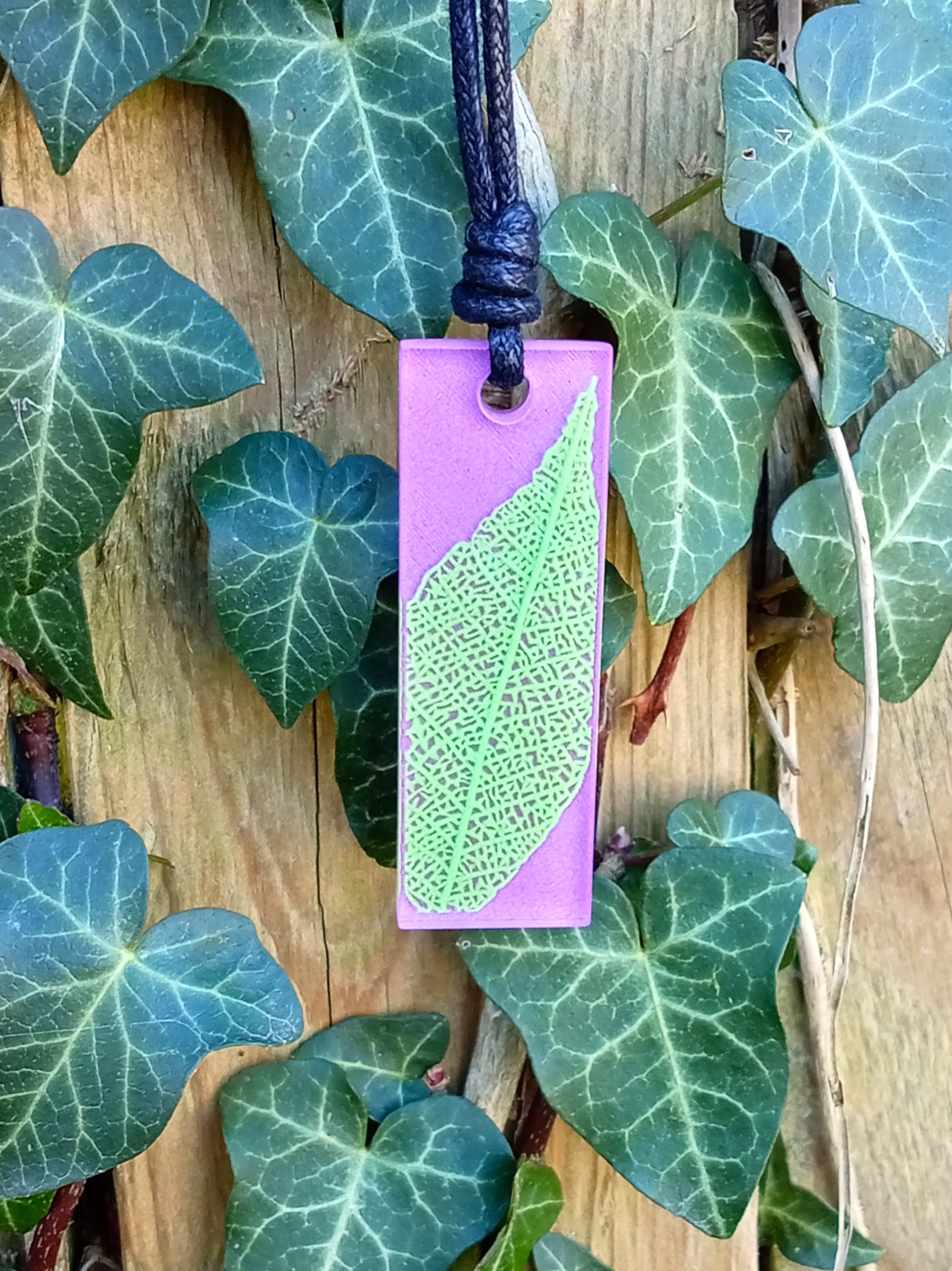 Purple rectangular pendant with bright green leaf skeleton design against ivy covered wood in natural daylight
