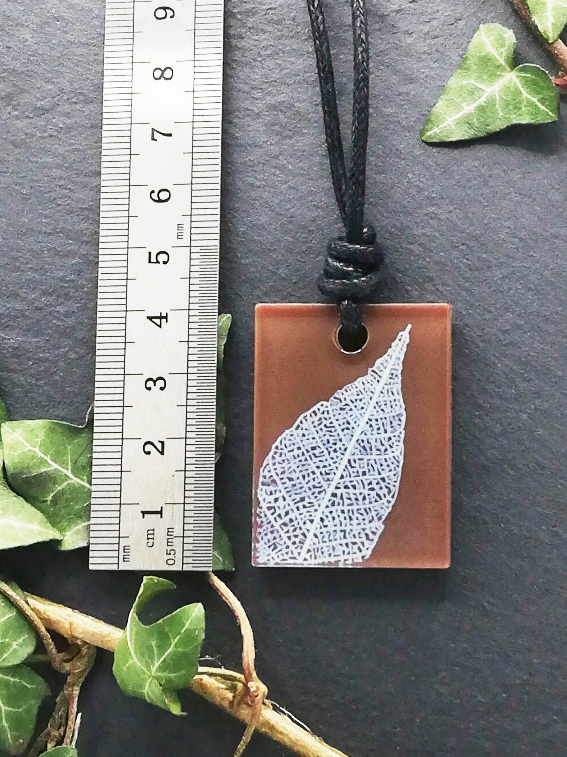 Warm brown rectangular pendant with cream leaf skeleton design shown next to a ruler displaying centimetres for scale