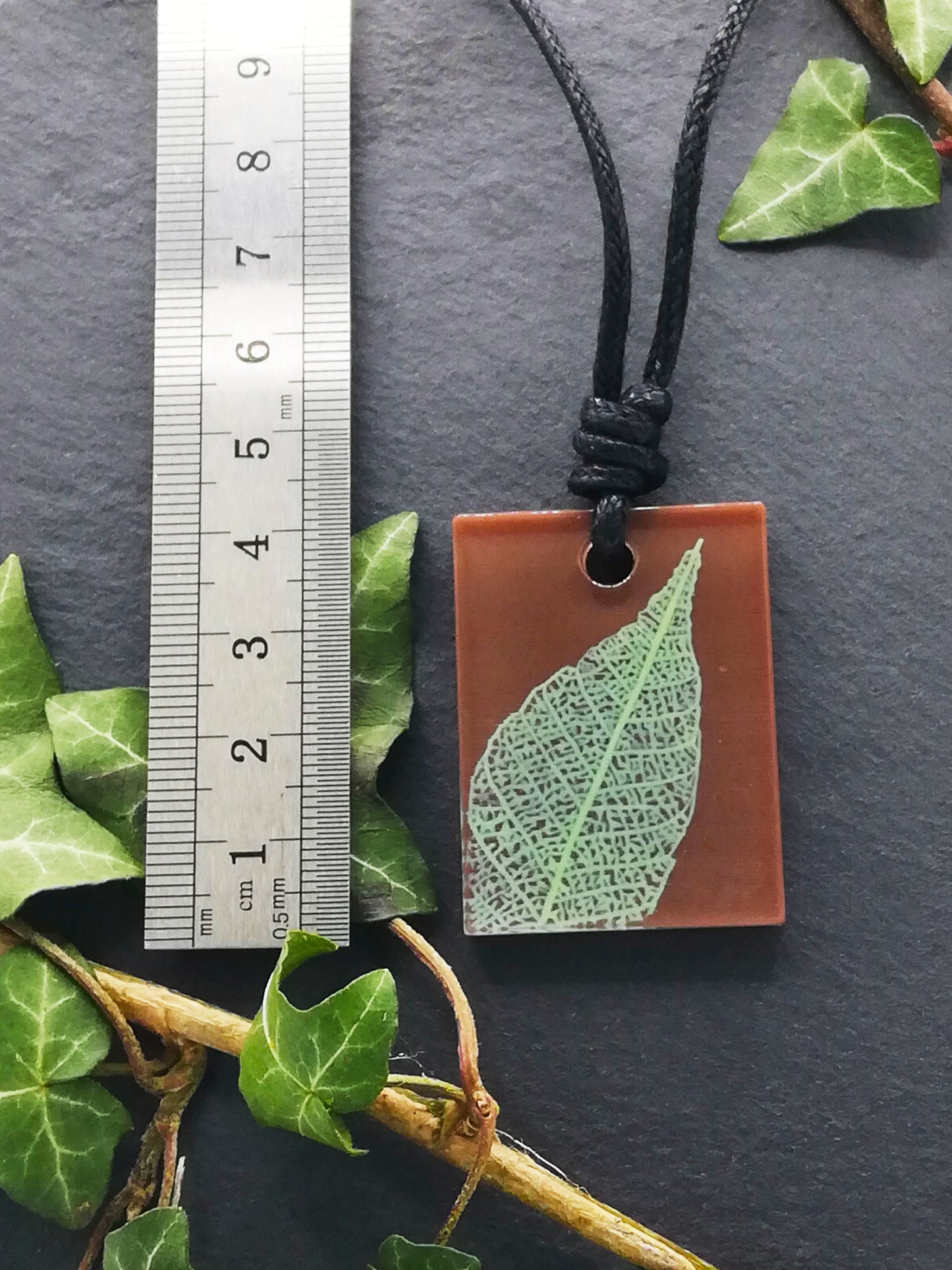 Warm brown rectangular pendant with pale green leaf skeleton design shown next to a ruler displaying centimetres for scale