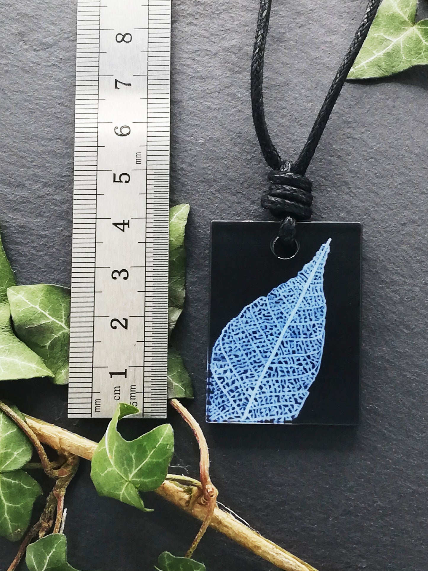 Black rectangular pendant with white leaf skeleton design shown next to a ruler displaying centimetres for scale