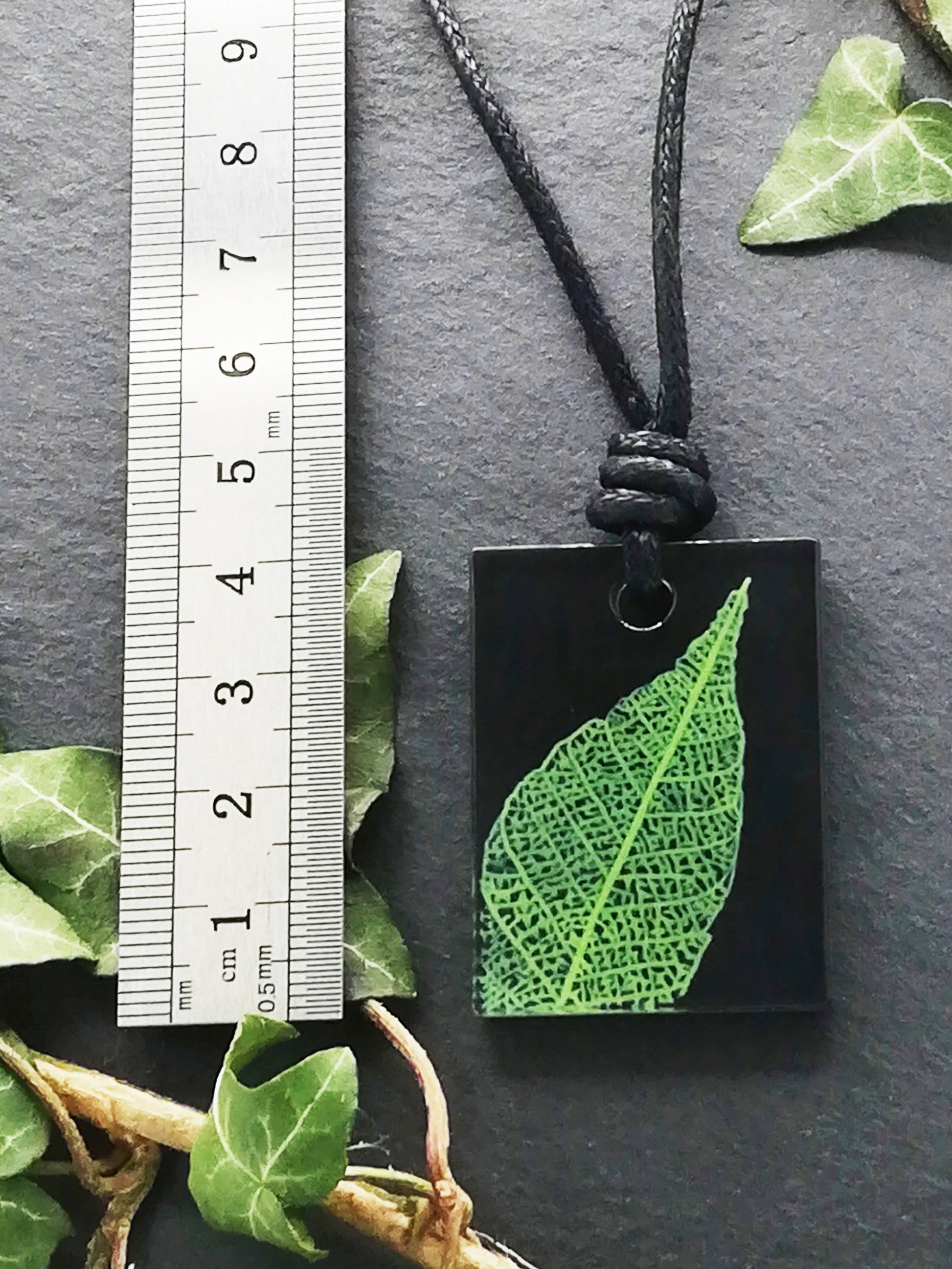 Black rectangular pendant with bright green leaf skeleton design shown next to a ruler displaying centimetres for scale