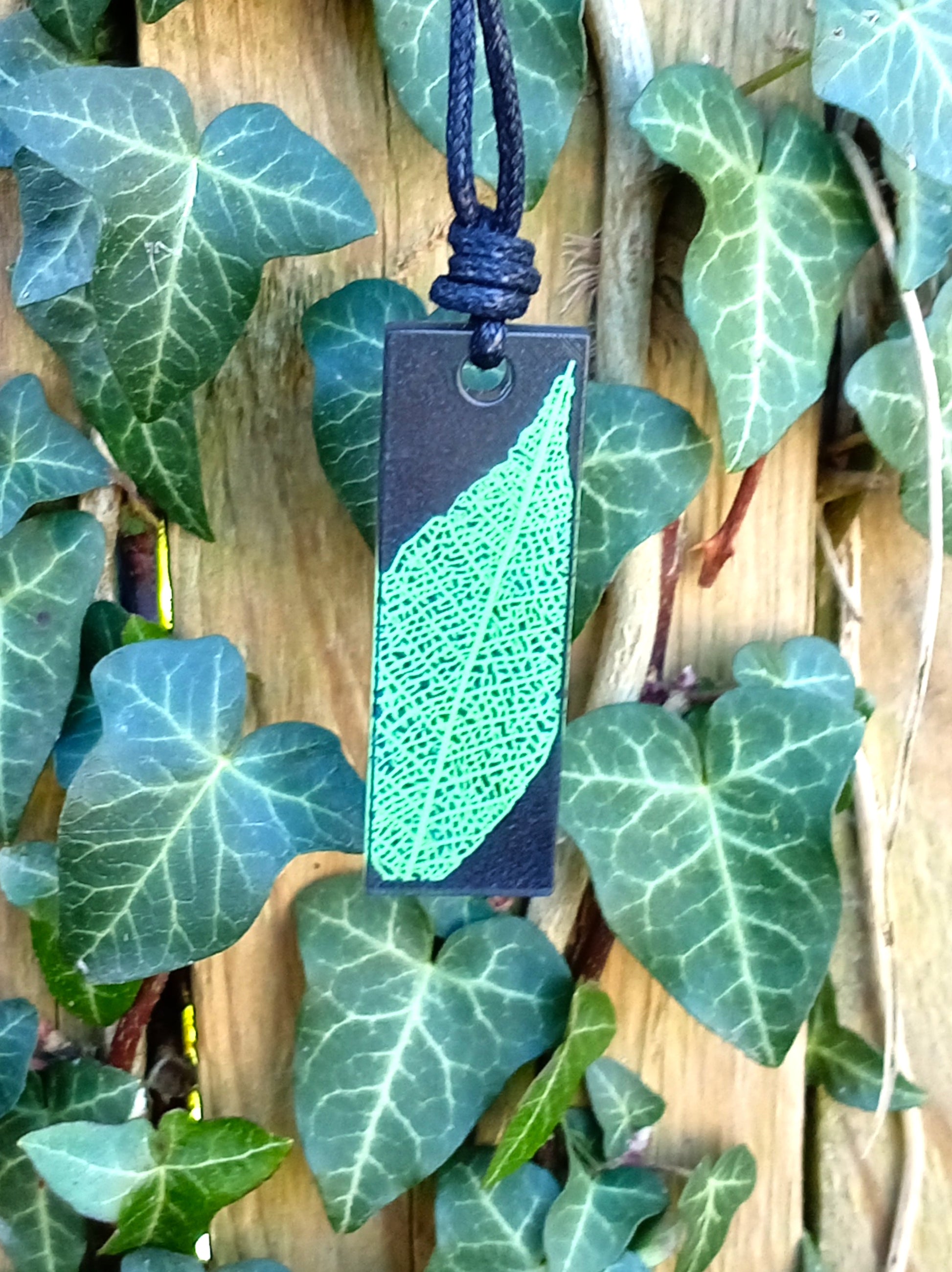 Black rectangular pendant with bright green leaf skeleton design against  ivy covered wood in natural daylight