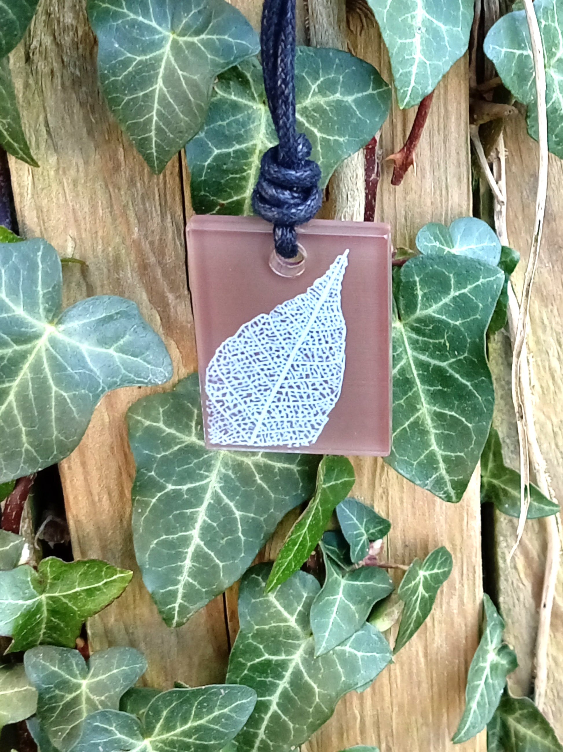 Warm brown rectangular pendant with cream leaf skeleton design against ivy covered wood in natural daylight