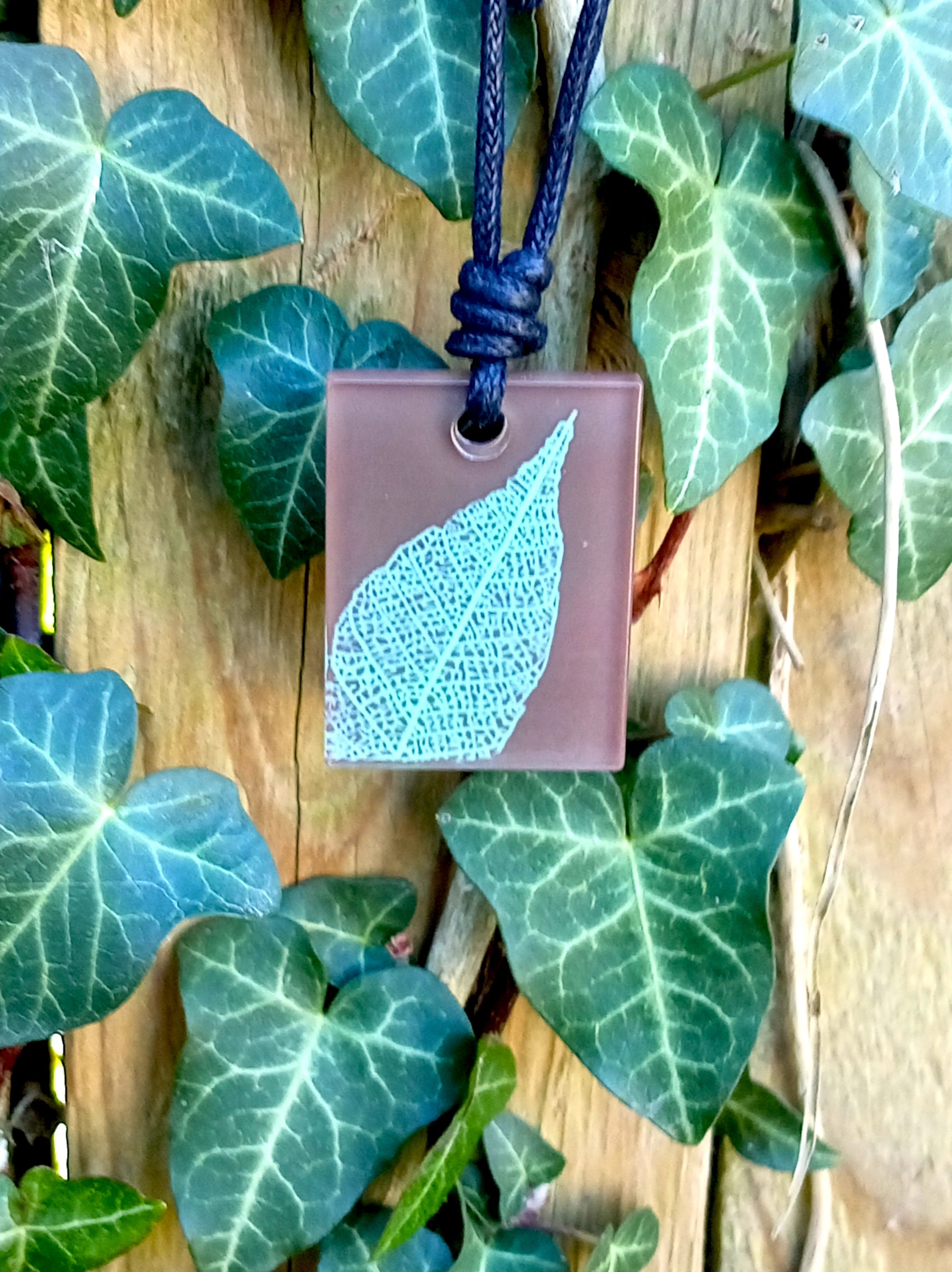 Warm brown rectangular pendant with pale green leaf skeleton design against ivy covered wood in natural daylight