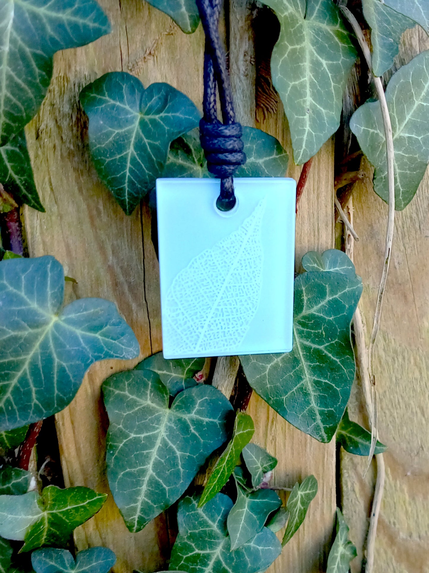 Light blue rectangular pendant with white leaf skeleton design against ivy covered wood in natural daylight