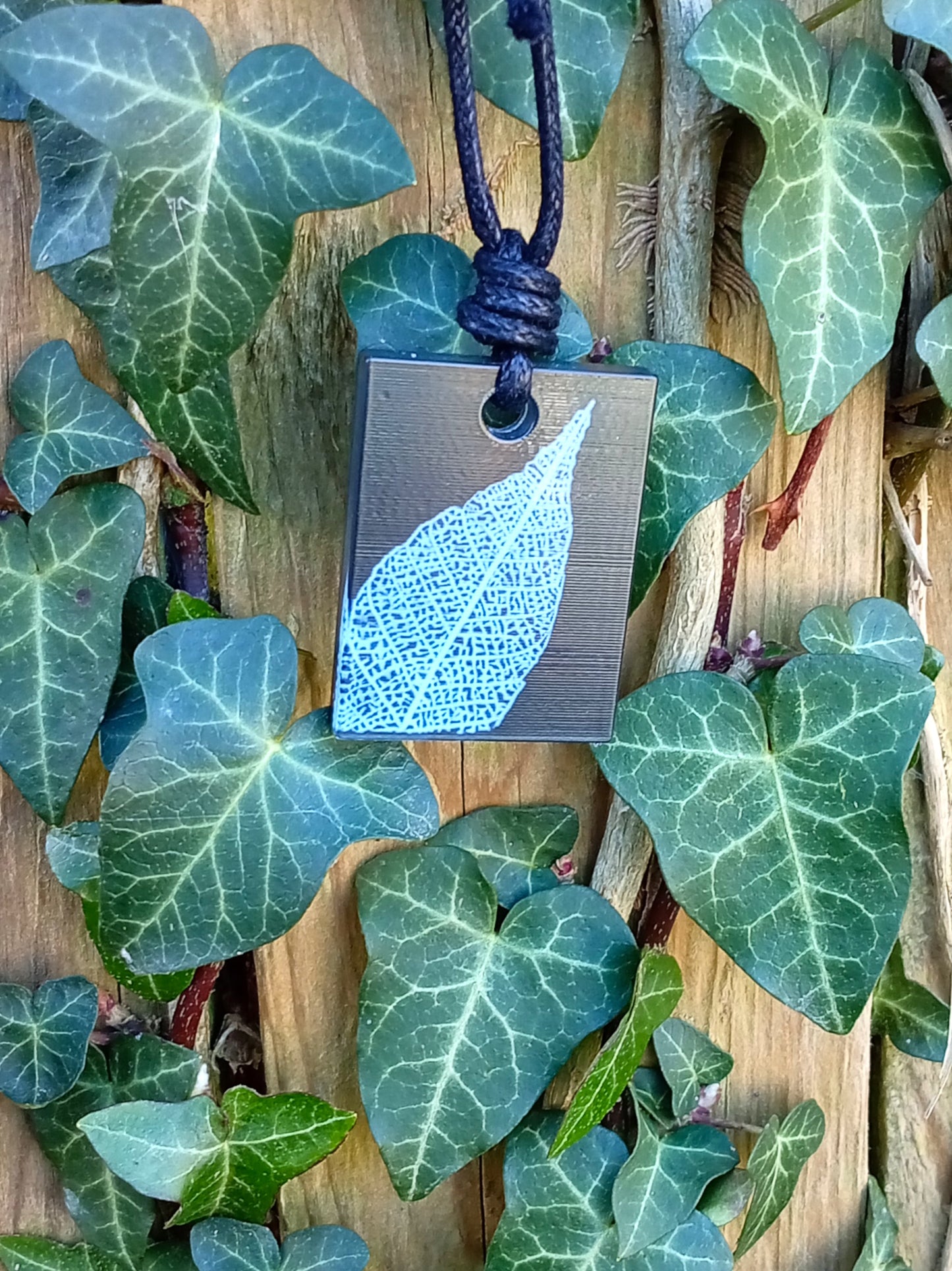 Black rectangular pendant with white leaf skeleton design against ivy covered wood in natural daylight