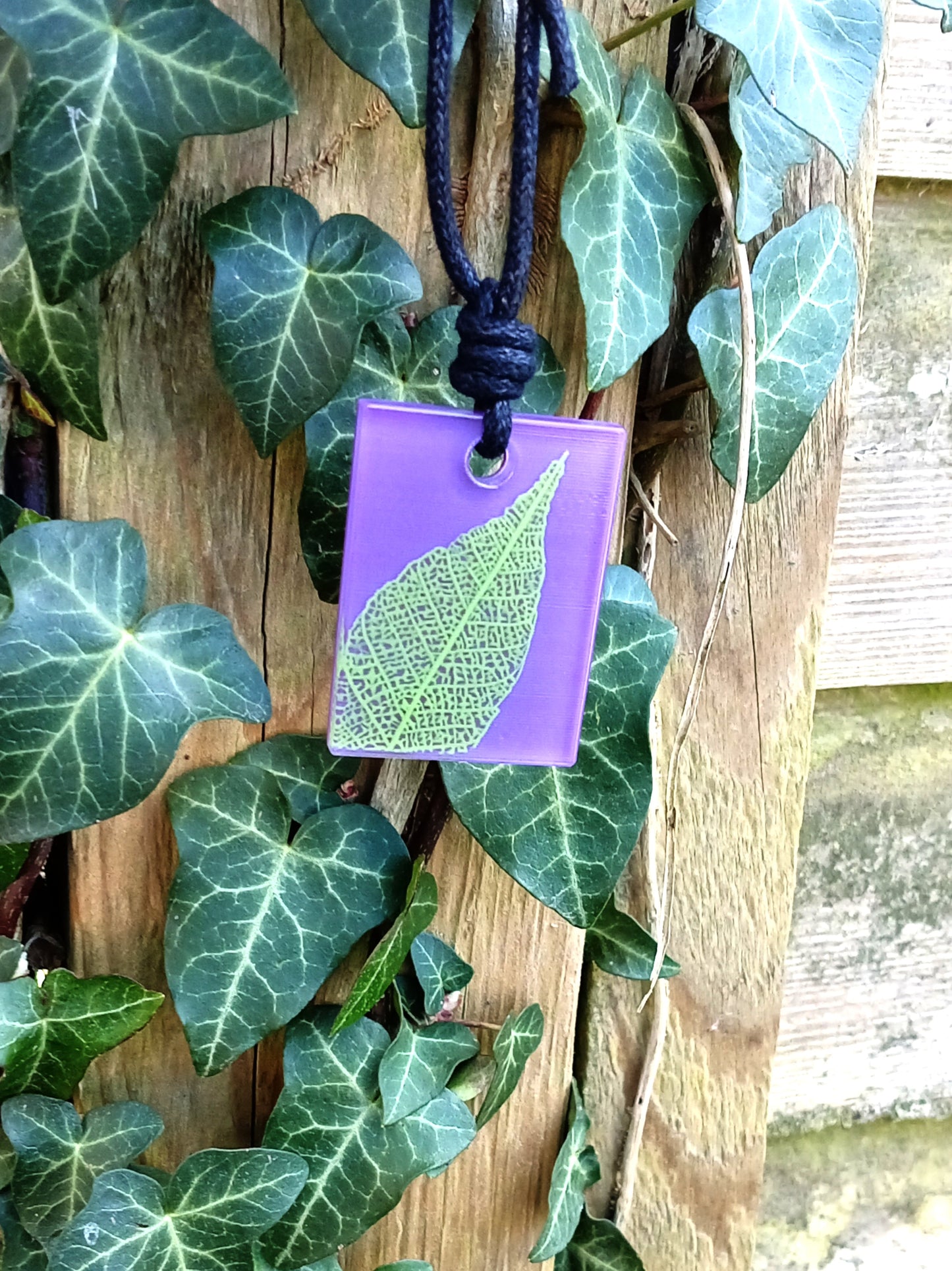 Purple rectangular pendant with bright green leaf skeleton design against ivy covered wood in natural daylight